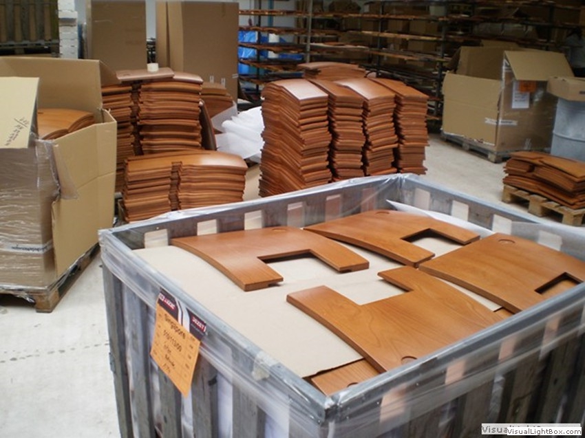 Braune Holzplatten mit Ausschnitten in einem Metallbehälter vor gestapelten Holzteilen.