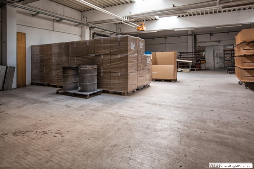 Große verpackte Stapel von Platten und Metallfässer auf Paletten in einer Werkhalle.
