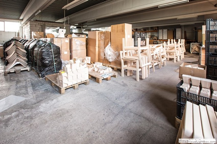 Verpackte Paletten, helle Stuhlgestelle und gestapelte Möbelteile in einer Lagerhalle.