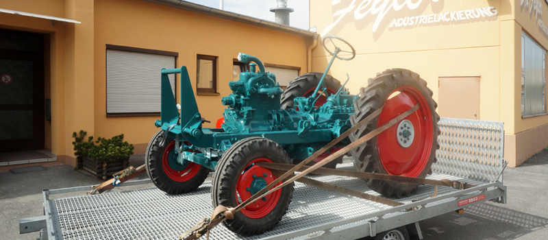 Türkisfarbener Traktor mit roten Rädern auf einem Anhänger vor einem Gebäude.